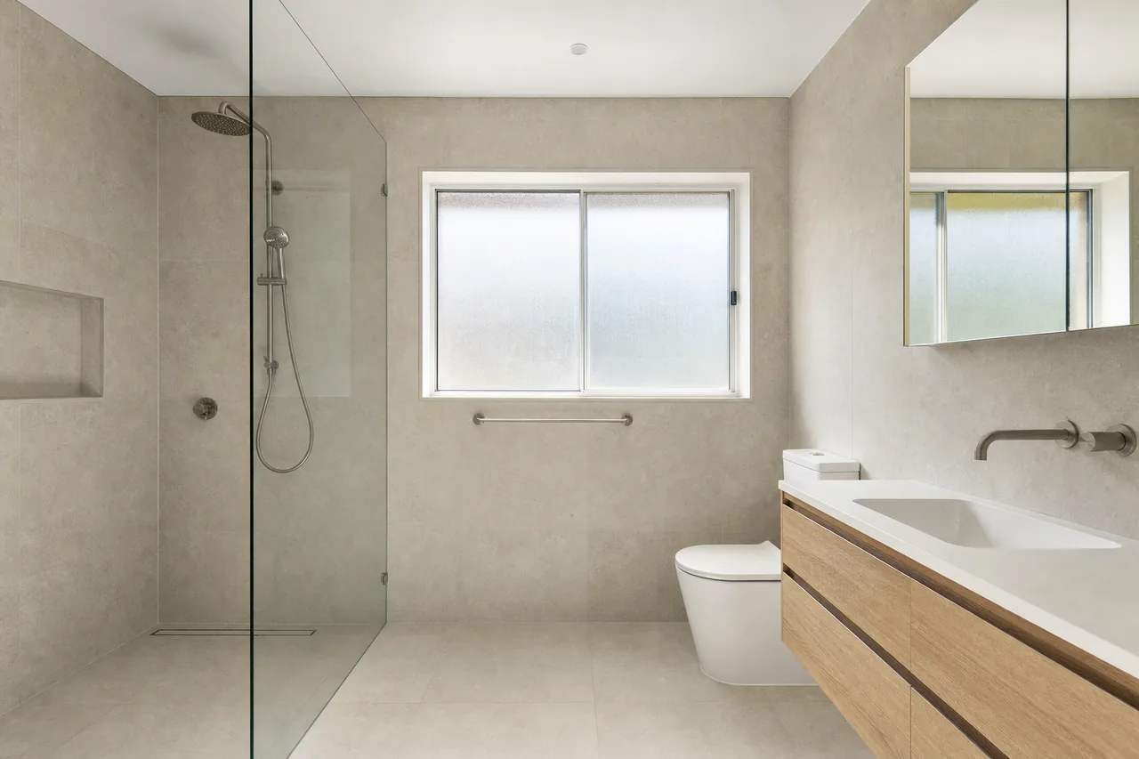 A finished bathroom with a frameless glass walk-in shower, large pale wall tiles, a frosted window and a wall-hung timber-look vanity — bathroom renovation, Newcastle and Lake Macquarie. Illustrative example.